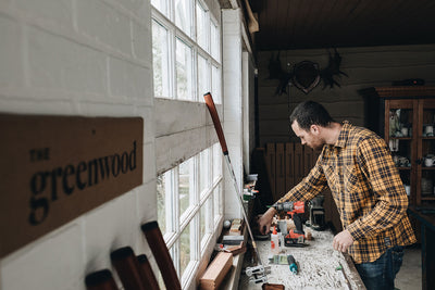 In wood shop where Greenwood handmade handcrafted solid wood golf putters are made, man in yellow plaid flannel in front of windows is reaching with his right arm over workbench where golf putter sits and sign to left reads the greenwood. 