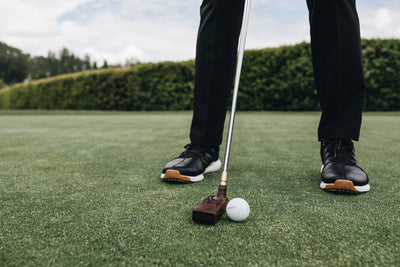 Solid wood putter in front of golfer's feet in black golf shoes. Handcrafted solid wood club head is resting on the putting green grass next to white golf ball.